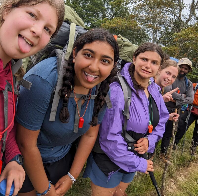 A group of six young hikers poses for a photo on a trail. They are wearing backpacks and hiking gear, and some are sticking their tongues out playfully. The hikers appear to be enjoying their time outdoors, surrounded by trees and greenery. The photo captures a moment of camaraderie and fun during their hiking adventure.